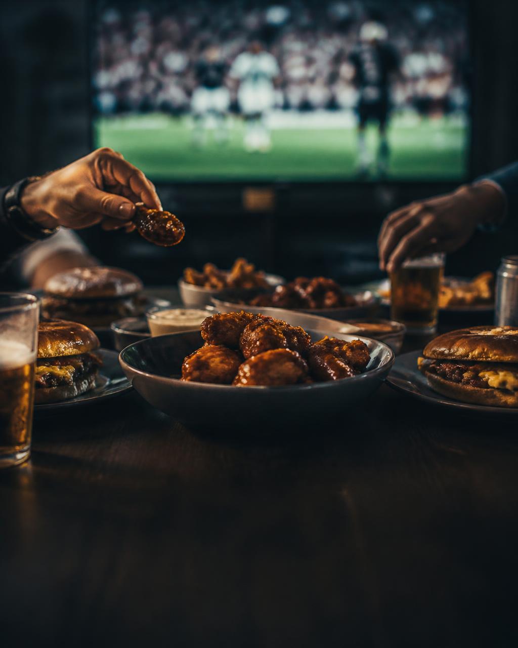 A loaded game-day table of wings, burgers and drinks with hands reaching in